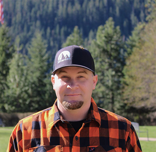 A grinning with a short beard wearing a baseball cap and an orange and black plaid flannel button down shirt.