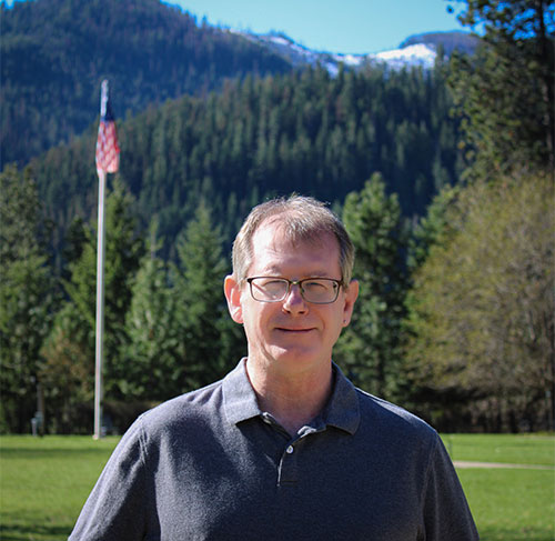 A smiling man wearing glasses and a grey polo shirt.