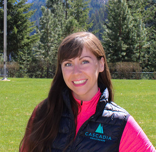 A smiling brunette woman wearing a bright pink turtleneck shirt and a blue down-filled vest.